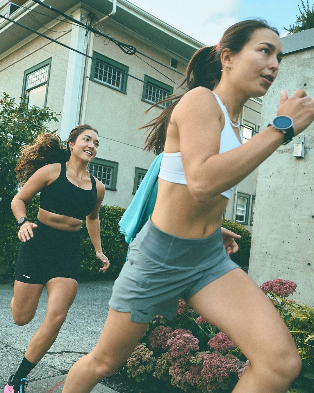 Two women in athletic wear are running outdoors on a paved path near a building, exercising together on a sunny day.