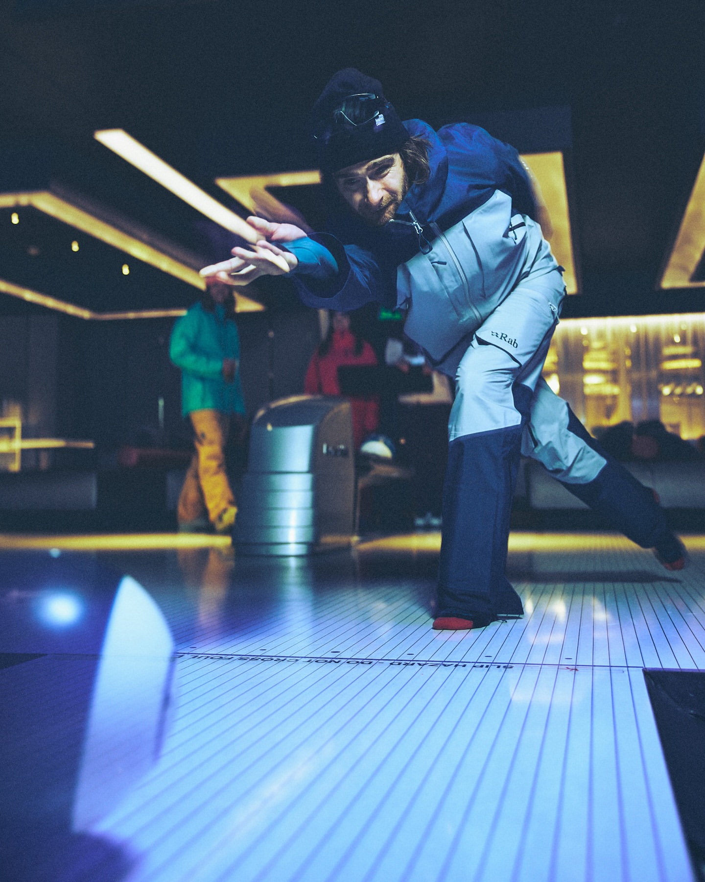 A man bowling indoors, wearing a blue and grey jacket and dark pants, is throwing a ball down a white lane, with people in the background.