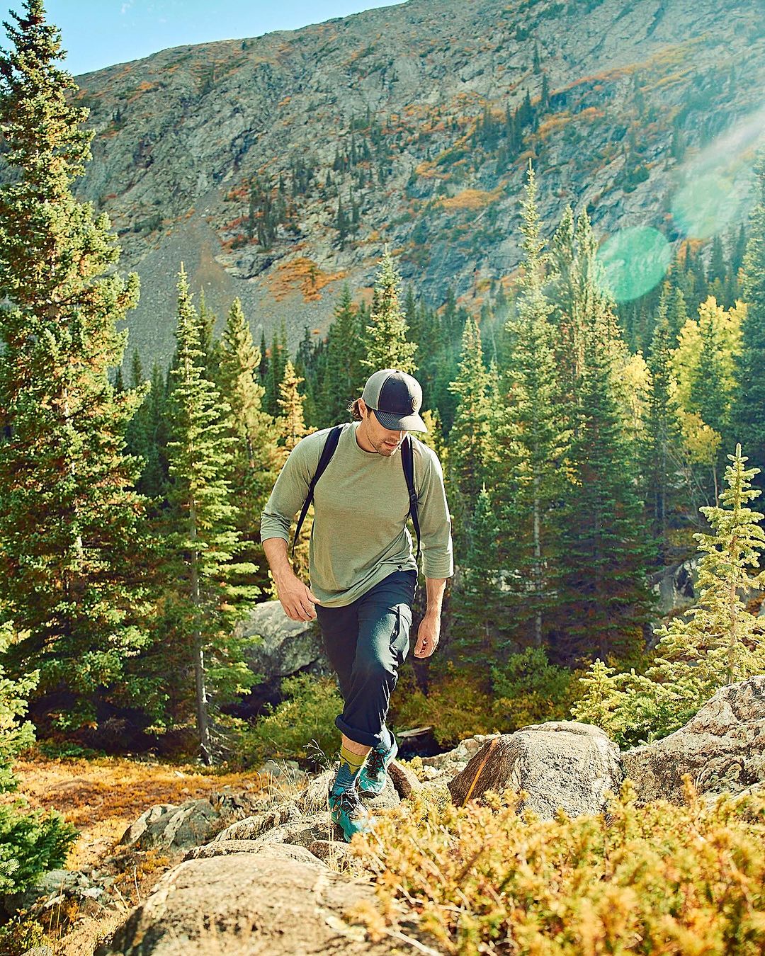 Man hiking uphill in a forest, surrounded by green trees and a mountain backdrop, wearing a long-sleeve shirt, dark pants, and hiking boots.
