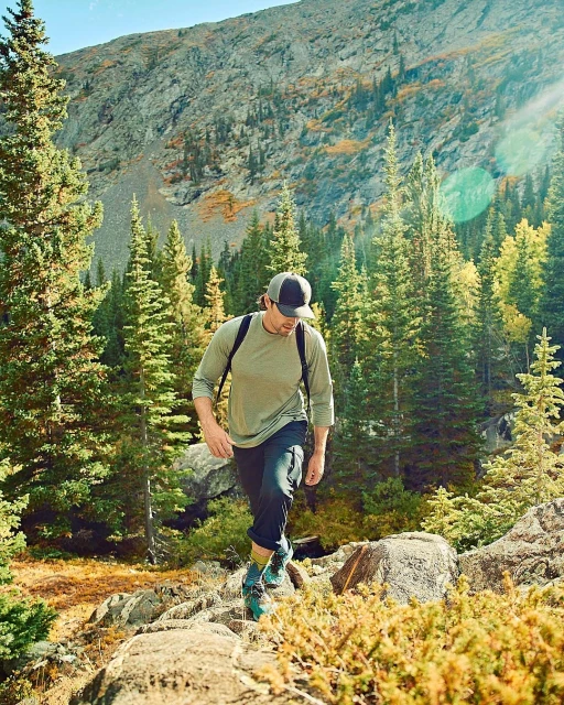 Man hiking uphill in a forest, surrounded by green trees and a mountain backdrop, wearing a long-sleeve shirt, dark pants, and hiking boots.