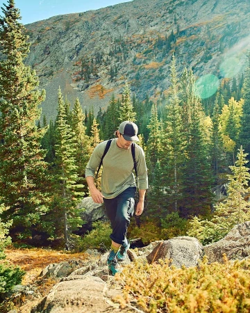 Man hiking uphill in a forest, surrounded by green trees and a mountain backdrop, wearing a long-sleeve shirt, dark pants, and hiking boots.