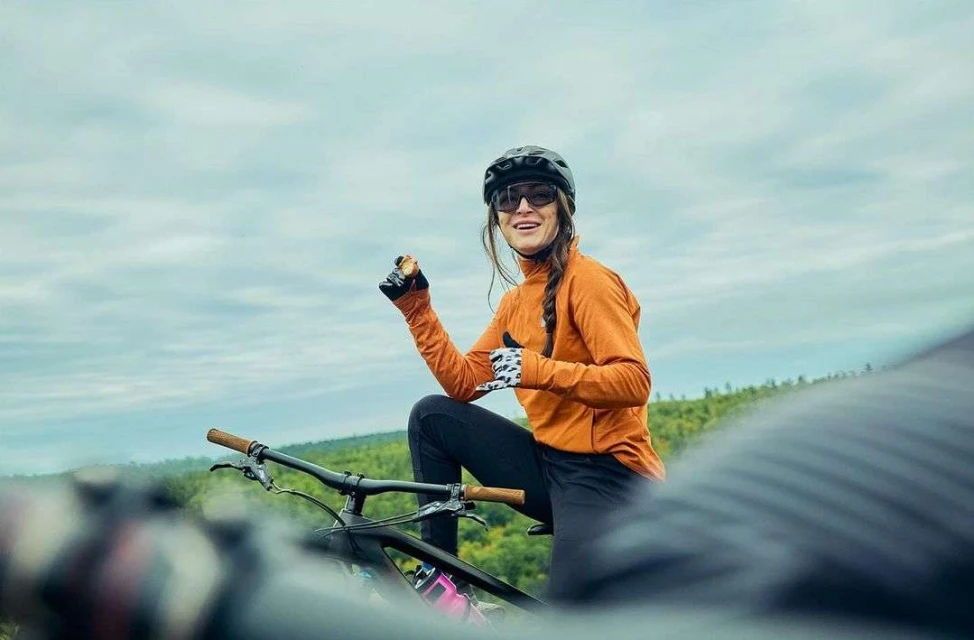 Adult female mountain biker in orange jacket pauses on a mountain trail to enjoy a snack with forest and cloudy sky in background.