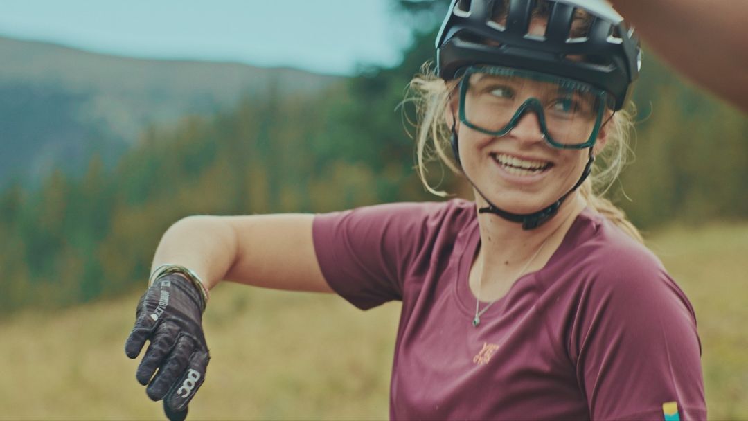 A smiling woman in sports goggles and a helmet looks off-camera while wearing a maroon athletic shirt, with a blurred outdoor background.