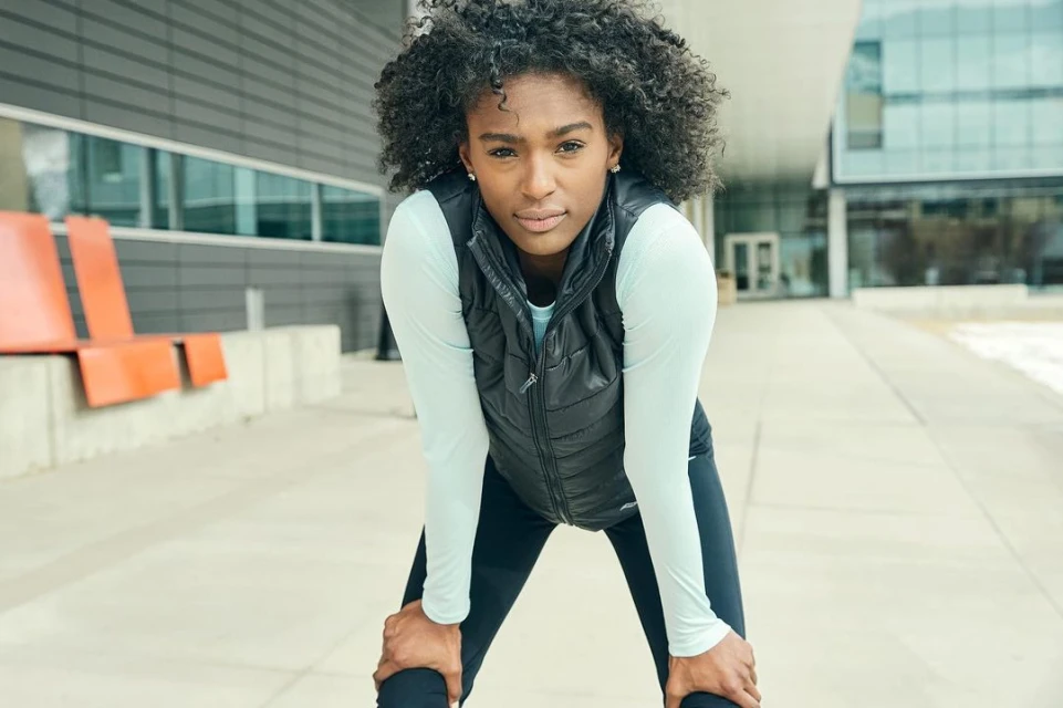 Adult woman rests with hands on knees outdoors in workout gear. Modern building and light pavement in the background.