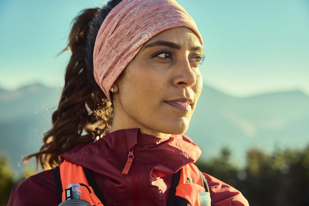 Woman in athletic wear with mountains in the background. The woman is wearing a headband and hydration pack, looking off into the distance.