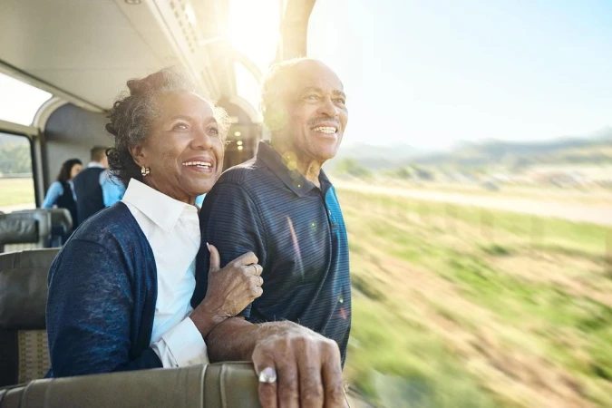 Smiling older couple enjoys a train ride, looking out the window at the blurred countryside, creating a sense of travel and adventure.