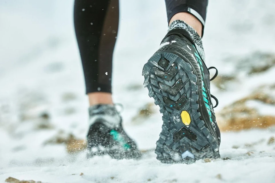 Low angle view of the sole of a black running shoe with snow cleats on the ground, with snow and another shoe behind.