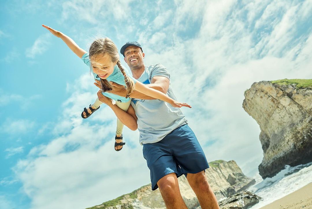 A dad holding his daughter with arms outstretched, simulating flight on a beach with ocean, rock formations, and partly cloudy sky.