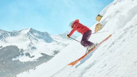 Skier in a red jacket and purple trousers carving down a steep snowy mountain slope under a clear blue sky
