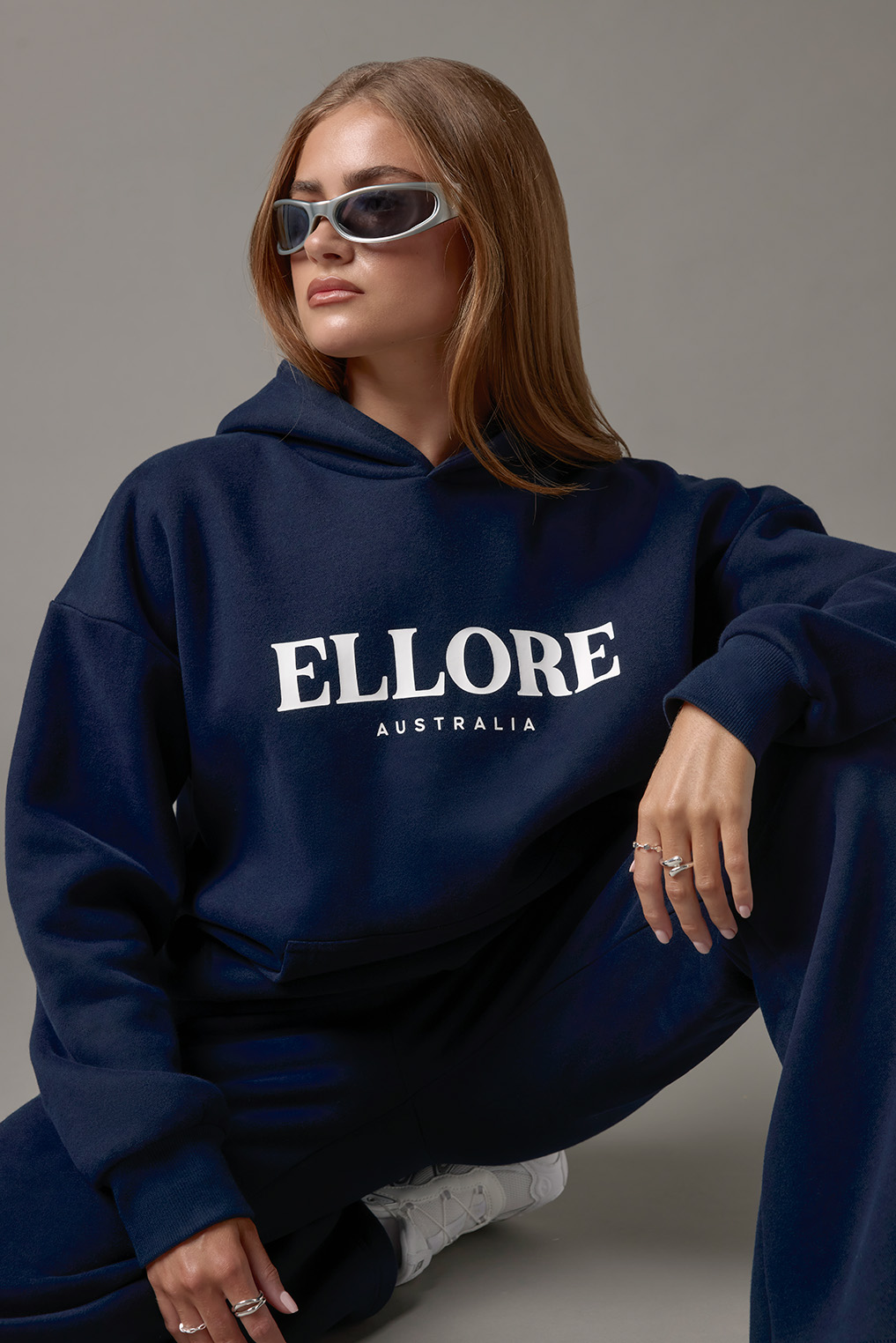 Woman poses in studio wearing oversized navy hoodie and pants. Visible text includes 'ELLORE AUSTRALIA'.