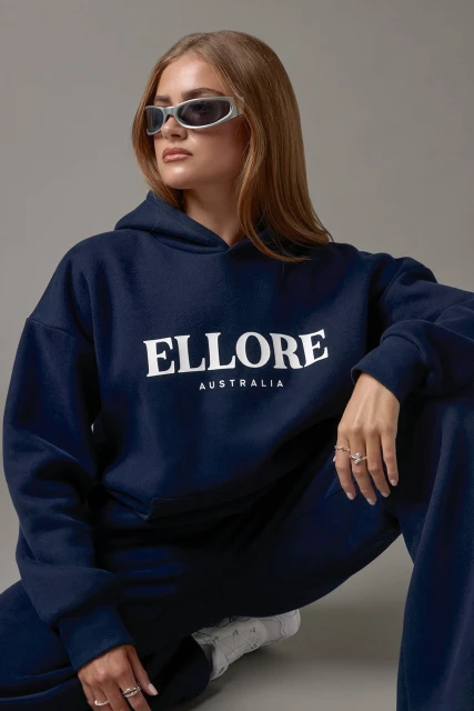 Woman poses in studio wearing oversized navy hoodie and pants. Visible text includes 'ELLORE AUSTRALIA'.