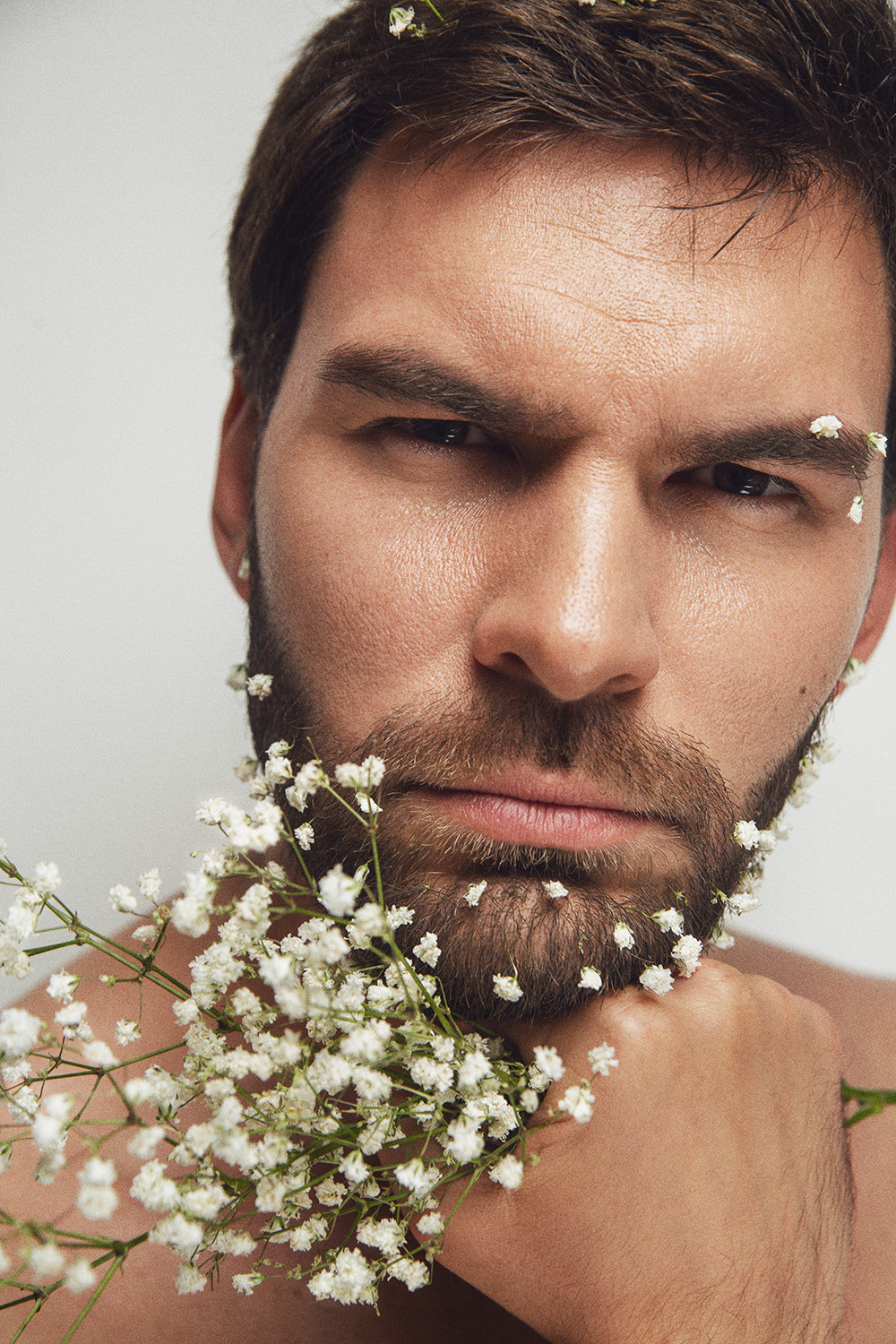 Portrait of a bearded adult male with small white flowers on his face and beard, holding a bouquet of flowers against a plain background.