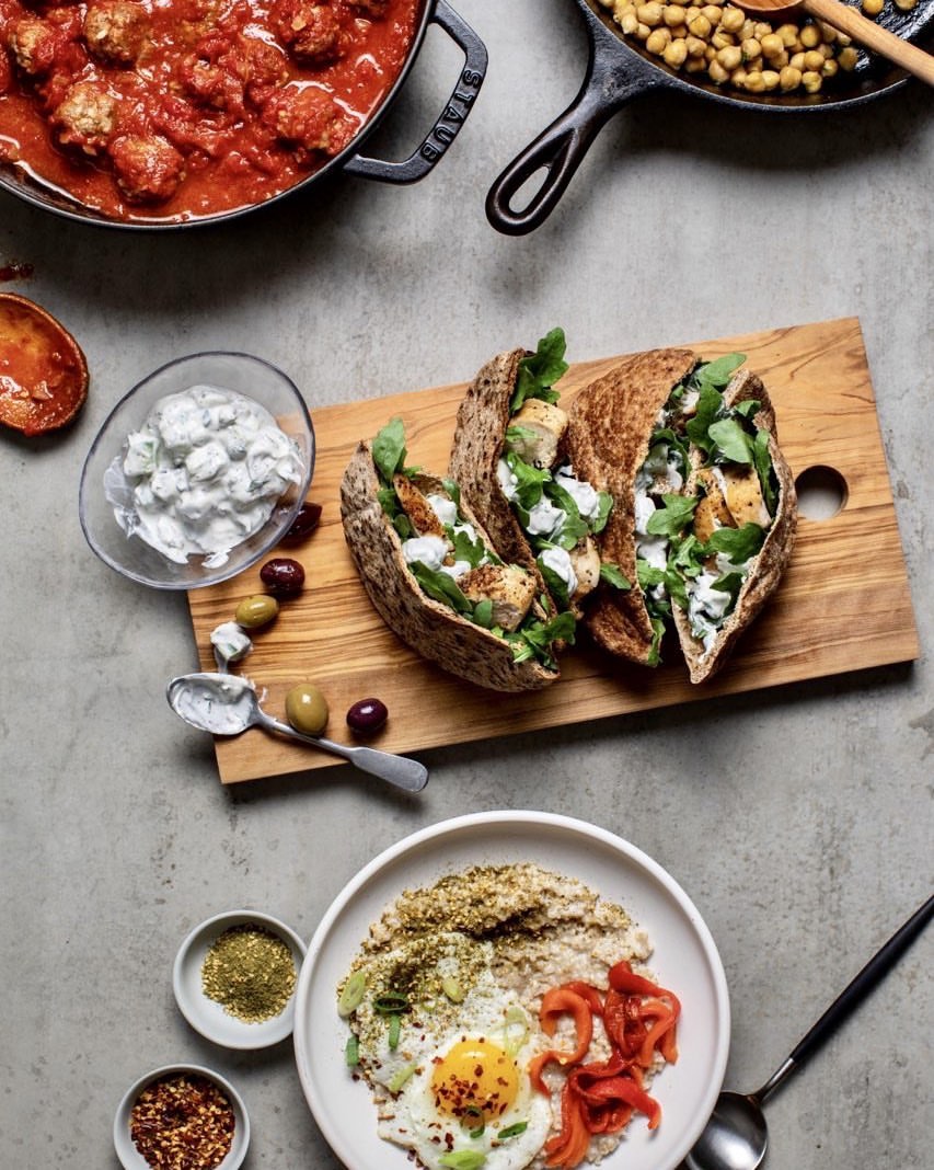 Overhead shot of chicken pita sandwiches, bowl with egg and roasted red peppers, bowl of tomato sauce, and garnishes on a gray surface.