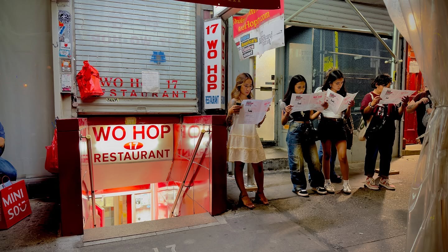 Four women standing in an urban setting are reading menus outside of the Wo Hop restaurant, with red signage and an entrance leading downstairs.