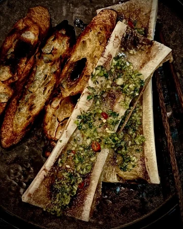 Overhead view of bone marrow dish featuring sliced baguettes and bone marrow topped with gremolata on a dark plate.