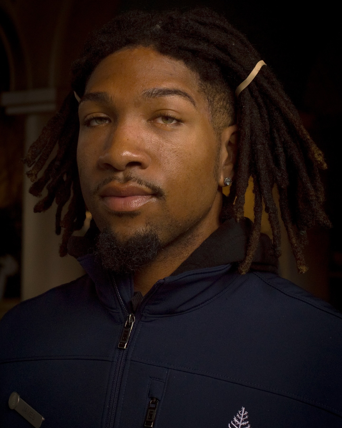 Close-up portrait of an adult male with dreadlocks wearing a dark jacket, looking directly at the camera with a neutral expression.