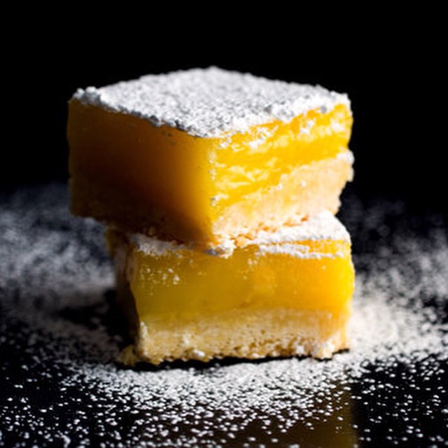 Stacked lemon bars dusted with powdered sugar on a black surface, close-up shot, dessert presentation, studio lighting, high angle.
