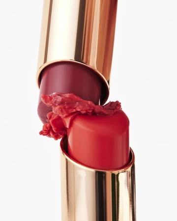 Macro shot of two broken golden lipstick tubes with smashed red and coral lipstick product smeared between them against white.