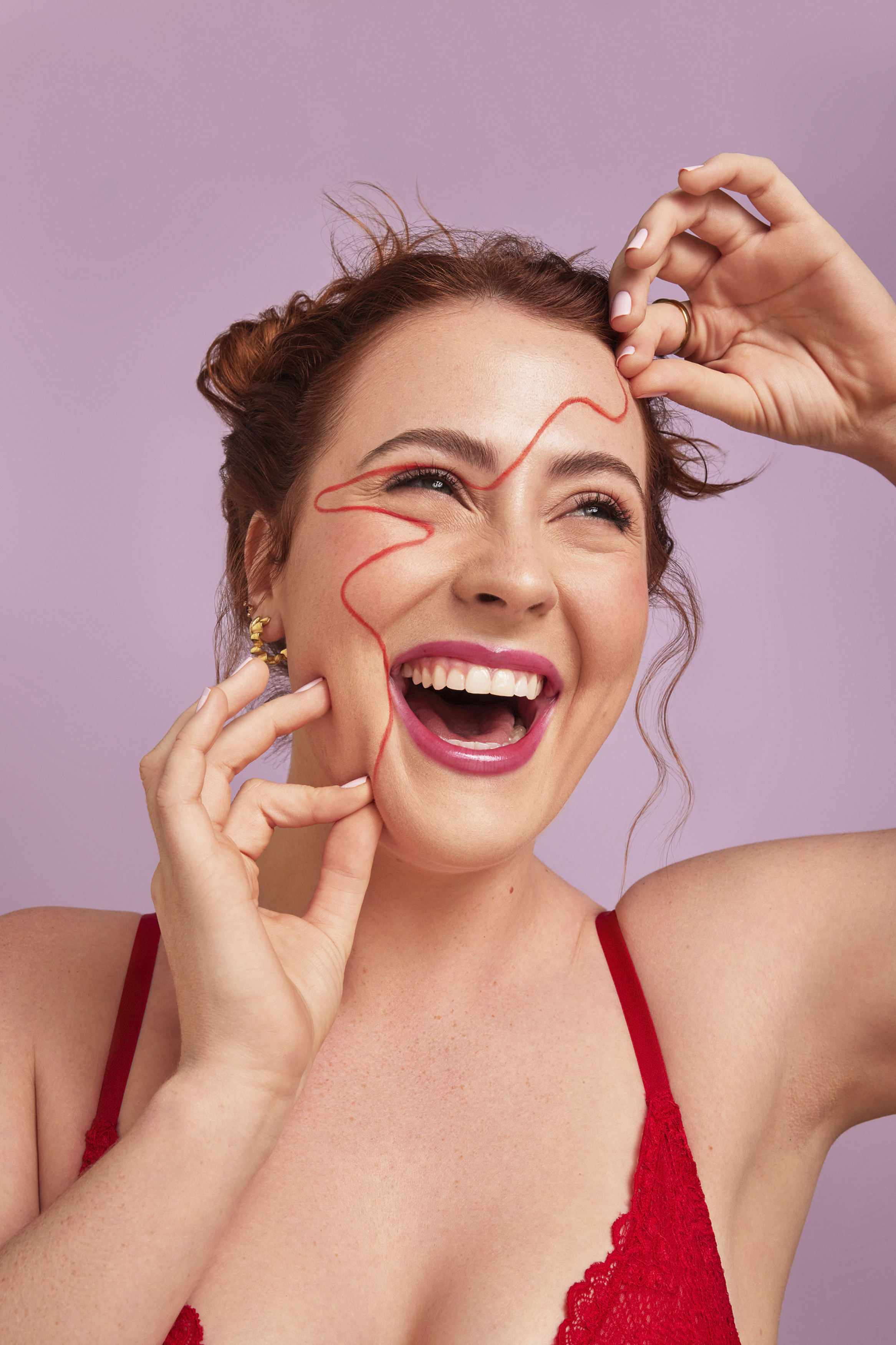 A laughing woman with red hair holds a wavy red thread across her face wearing a red lace top against a lilac studio backdrop.