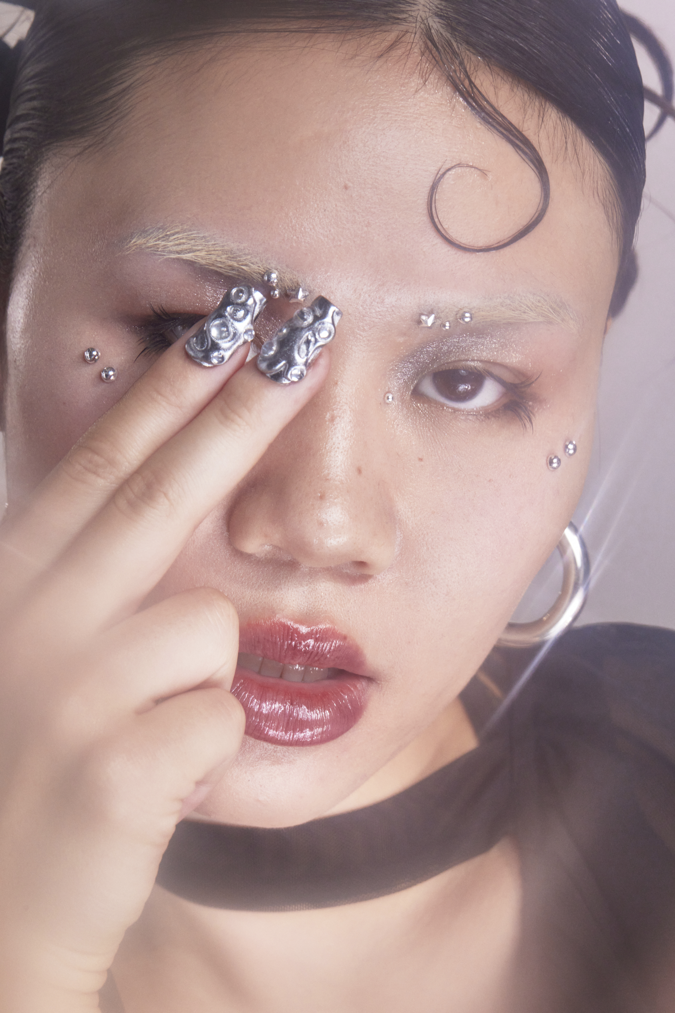 Close-up beauty portrait of an East Asian woman with metallic textured nails touching her face, silver piercings, and glossy lips.