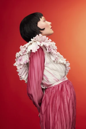 Profile portrait of a woman with dark bobbed hair wearing a pink ruffled top against a warm gradient background.