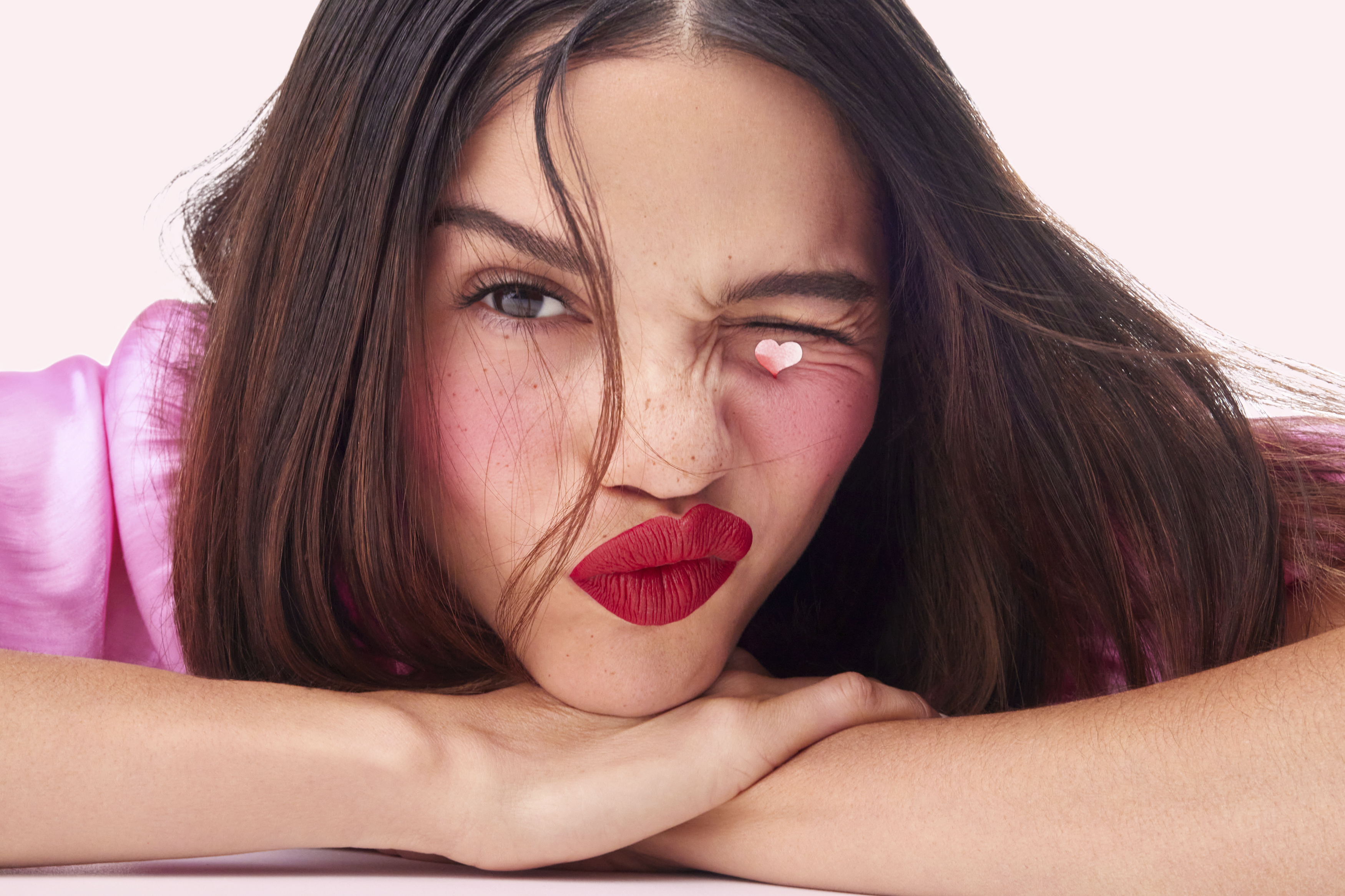 Close-up portrait of a woman winking with a tiny heart on her eyelid, wearing bright red lipstick and pink blush against a light background.
