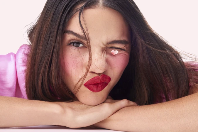 Close-up portrait of a woman winking with a tiny heart on her eyelid, wearing bright red lipstick and pink blush against a light background.