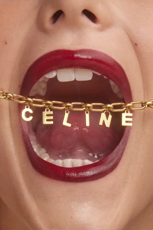 Extreme close-up of an open mouth with dark red lipstick holding a gold chain necklace with letter charms spelling CELINE.