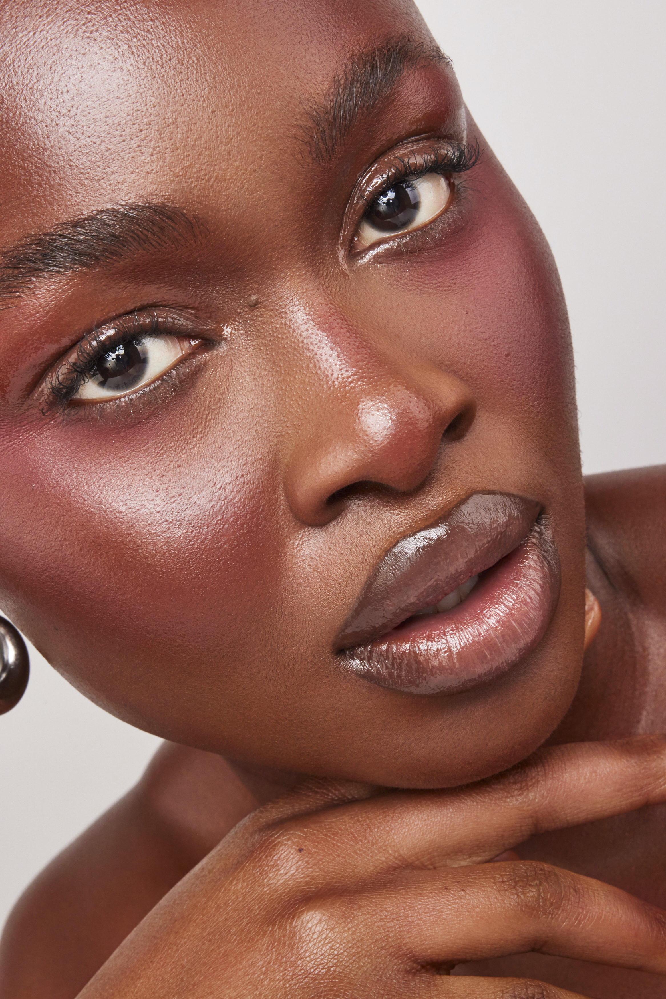 Close-up beauty portrait of a Black woman with dewy skin, glossy lips, and pink blush on a neutral background.