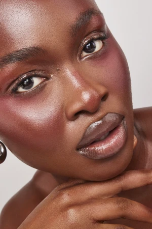 Close-up beauty portrait of a Black woman with dewy skin, glossy lips, and pink blush on a neutral background.