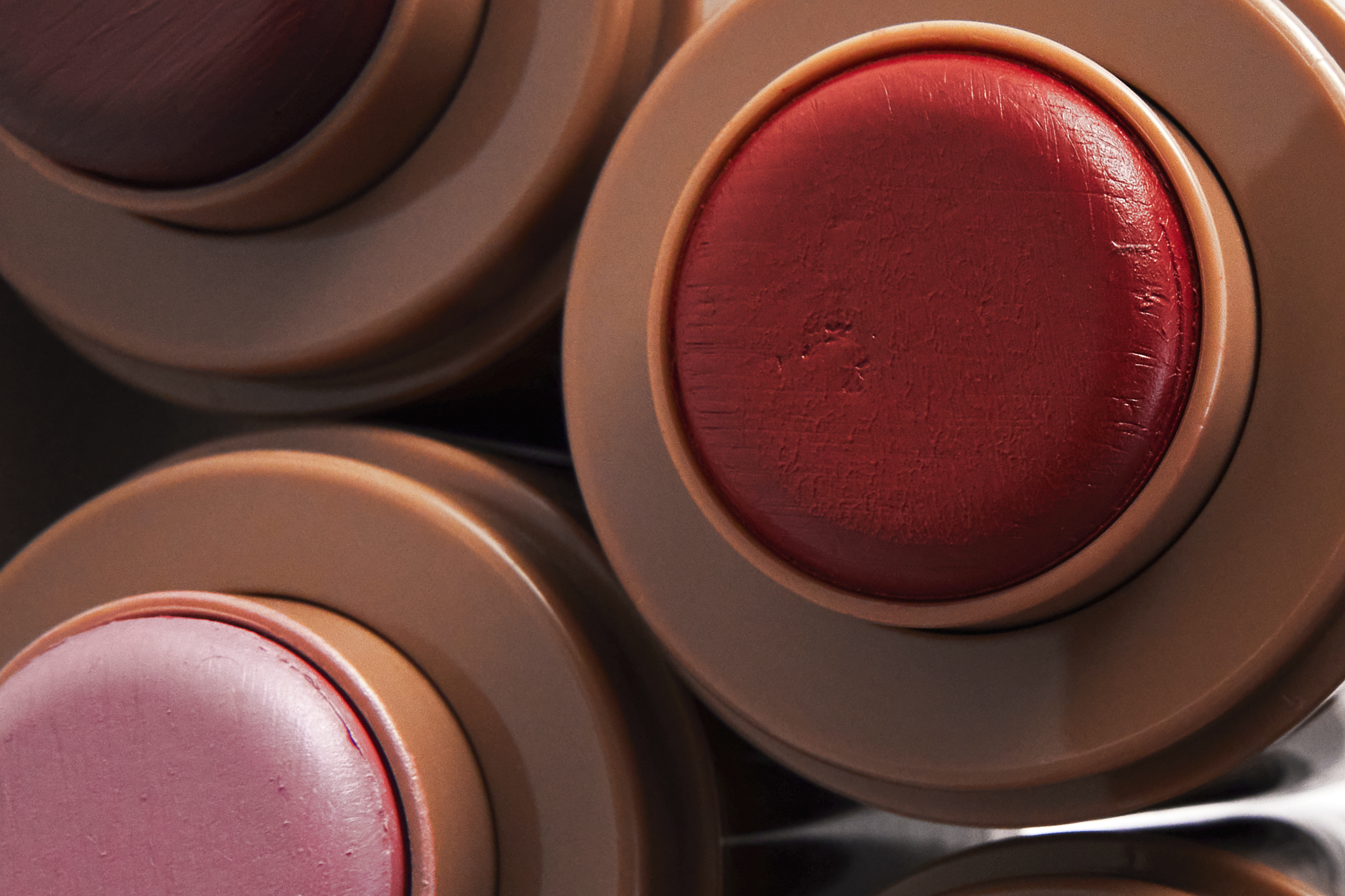 Top-down macro close-up of three cosmetic balm sticks in neutral beige plastic tubes showing red and rose shades.