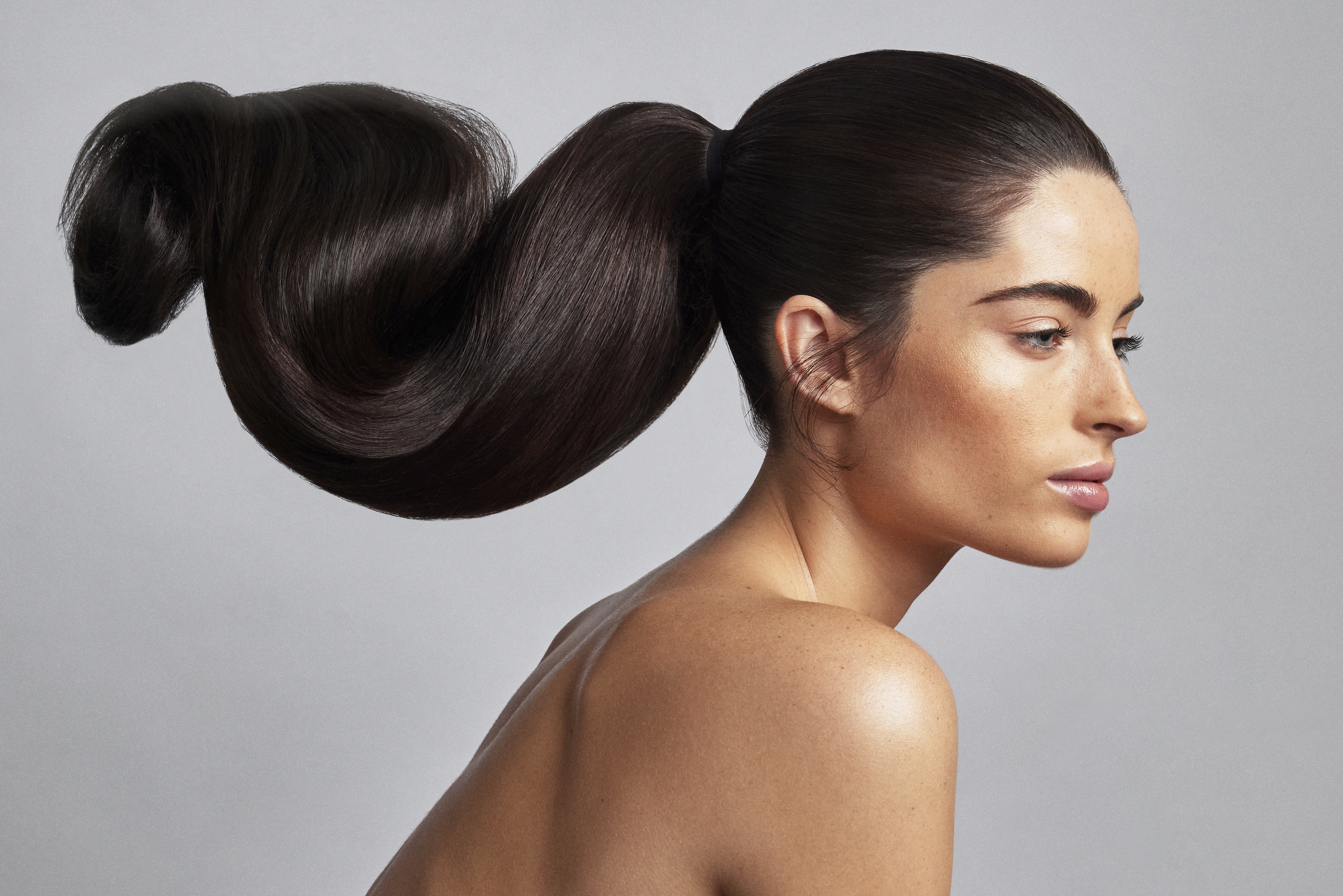 Profile portrait of a woman with dark brown hair styled in an elaborate sculptural ponytail against a neutral grey background.