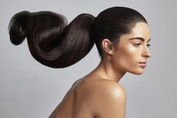 Profile portrait of a woman with dark brown hair styled in an elaborate sculptural ponytail against a neutral grey background.