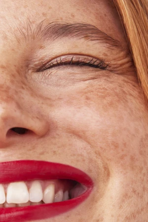 Extreme macro beauty portrait of a smiling woman with red lipstick and heavy freckles across her face.