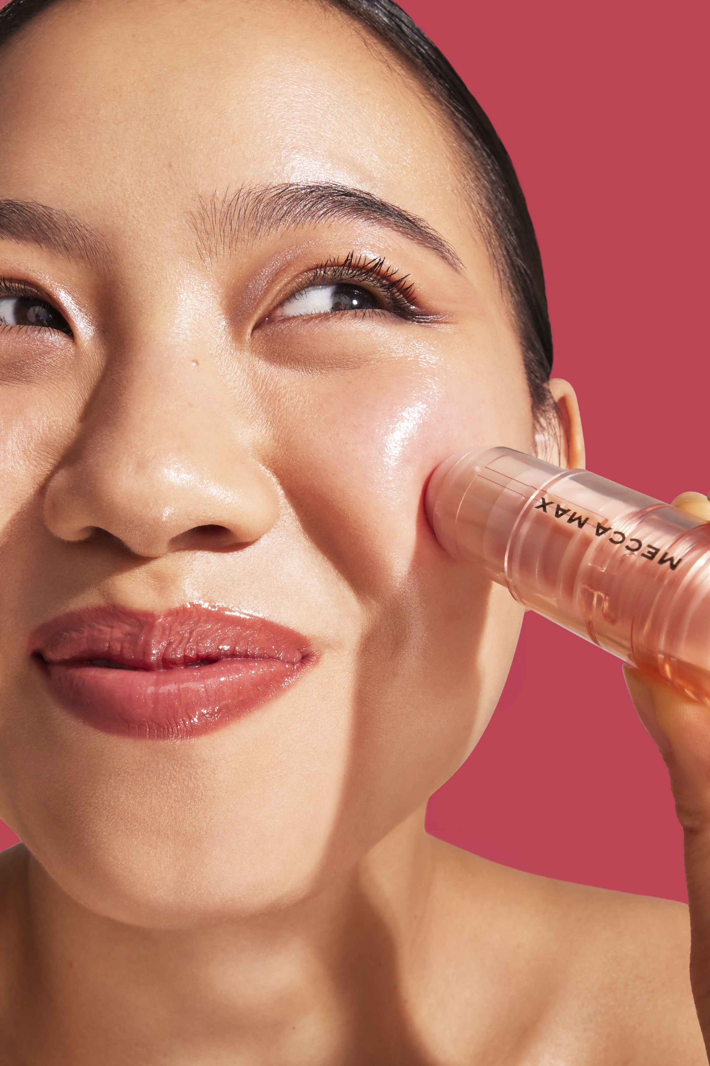 Close-up portrait of an East Asian woman applying a Mecca Max blush stick to her cheek against a vibrant pink background.