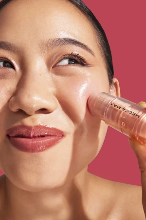 Close-up portrait of an East Asian woman applying a Mecca Max blush stick to her cheek against a vibrant pink background.