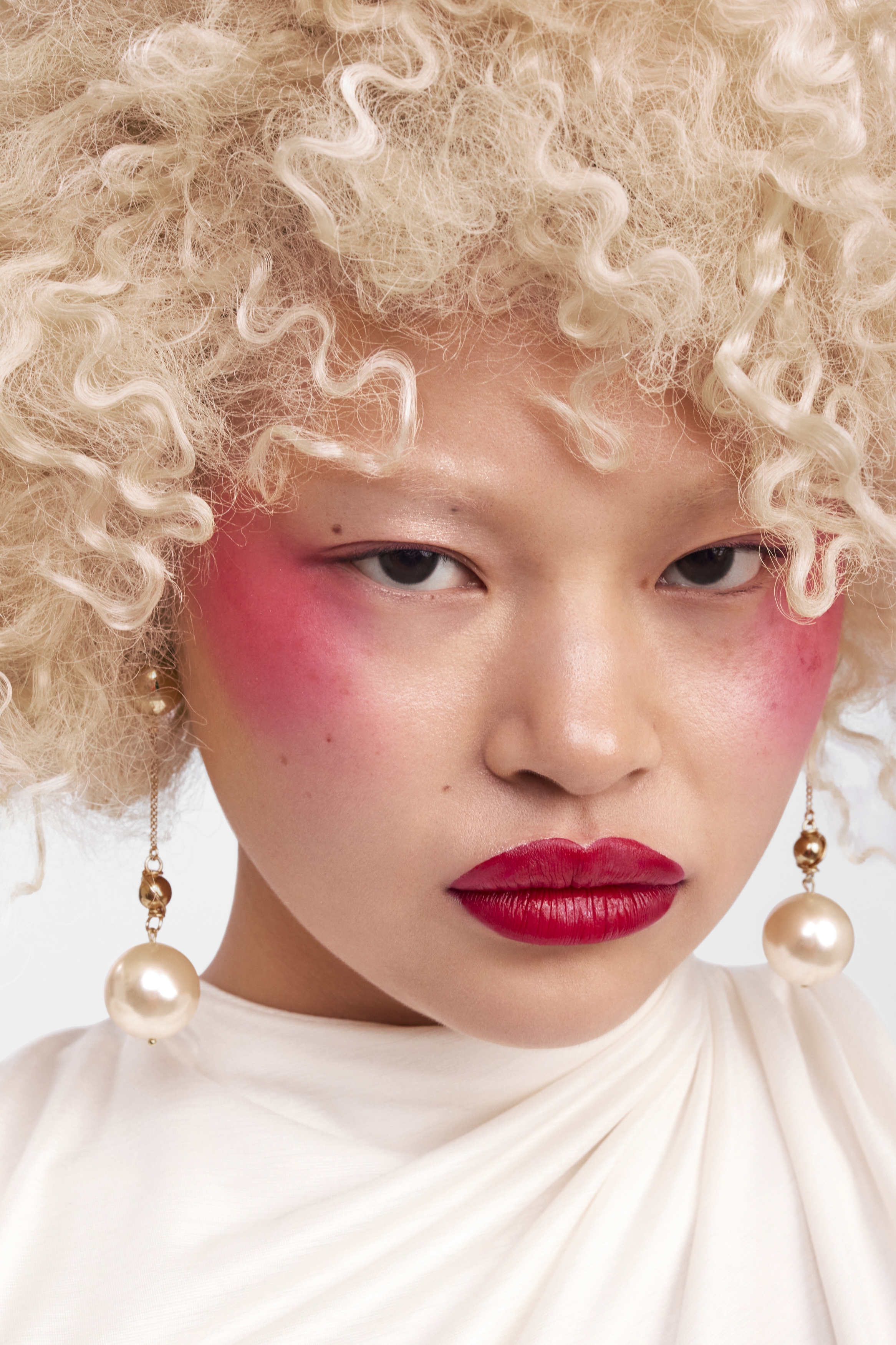 Close-up beauty portrait of an East Asian woman with curly blonde hair, vivid pink blush, red lipstick, and pearl drop earrings.