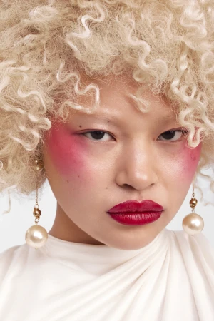 Close-up beauty portrait of an East Asian woman with curly blonde hair, vivid pink blush, red lipstick, and pearl drop earrings.