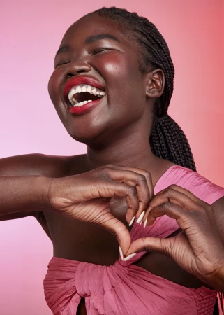 Joyful Black woman with braided hair making a heart shape with her hands while laughing, wearing red lipstick and a pink top.