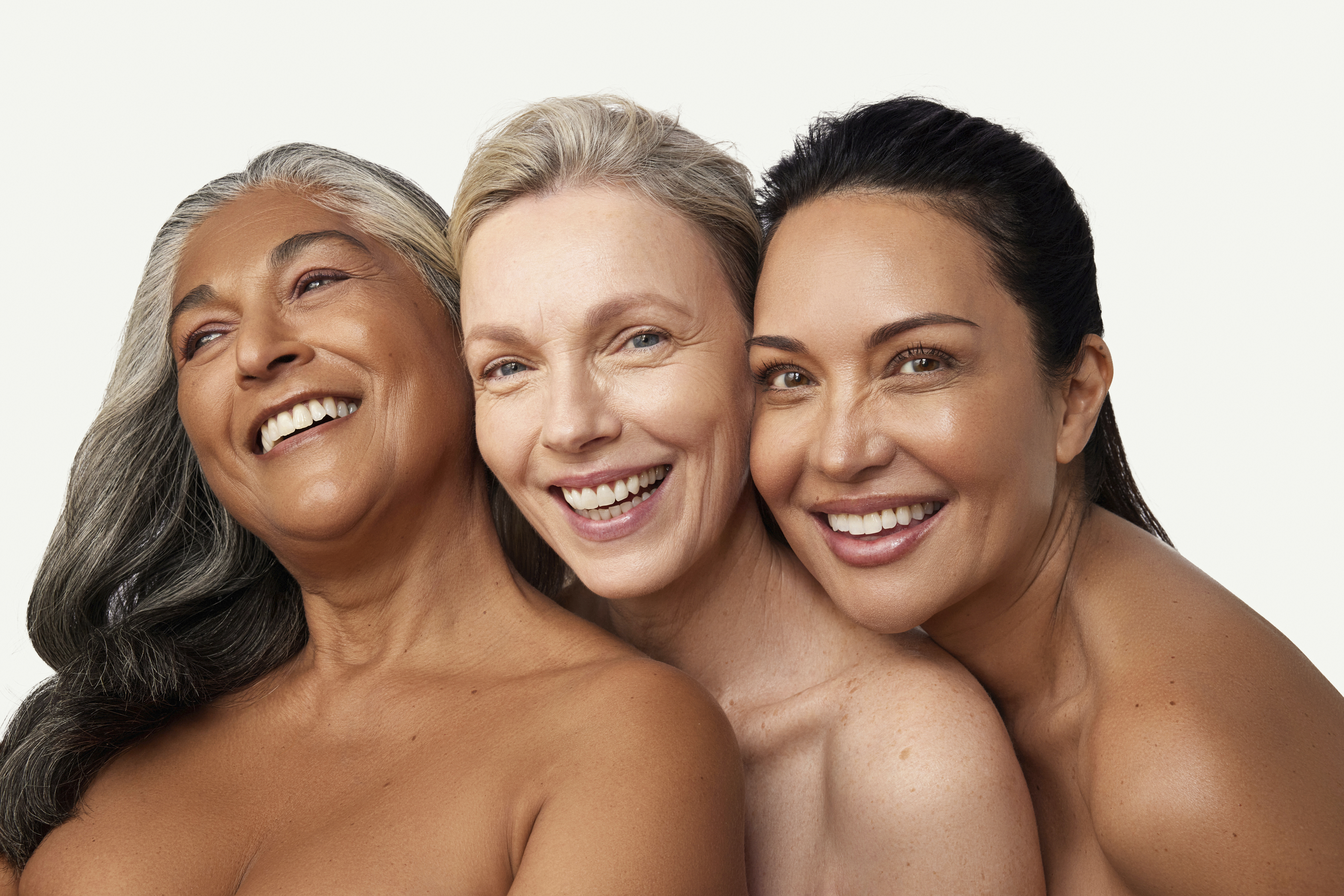 Three diverse women of different ages smiling together with bare shoulders against a white studio background.