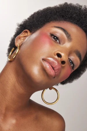 Close-up beauty portrait of a Black woman with short dark hair wearing thick gold hoop earrings and vibrant pink blush.