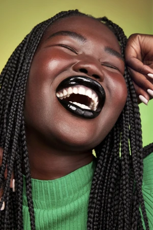 Close-up portrait of a Black woman with braided hair laughing, wearing glossy black lipstick and a green ribbed top against a green background