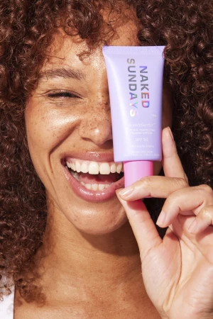Smiling woman with curly brown hair holding a purple tube of Naked Sundays sunscreen lotion over one eye in a beauty close-up.