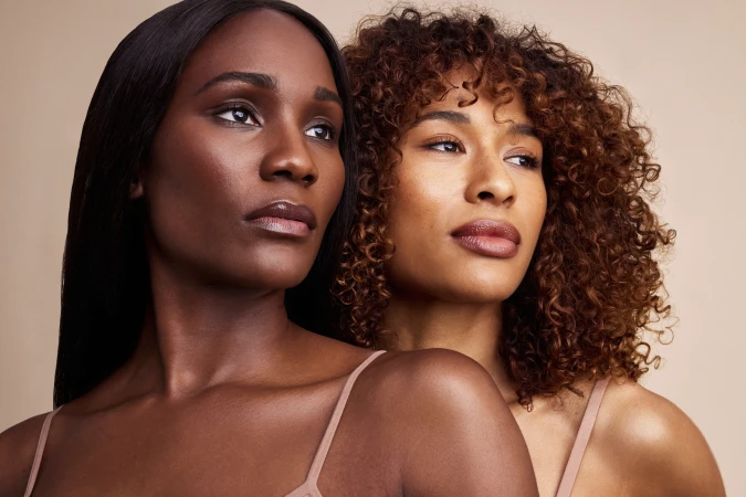 Close-up beauty portrait of two women with glowing skin and nude lip gloss against a warm beige studio backdrop.