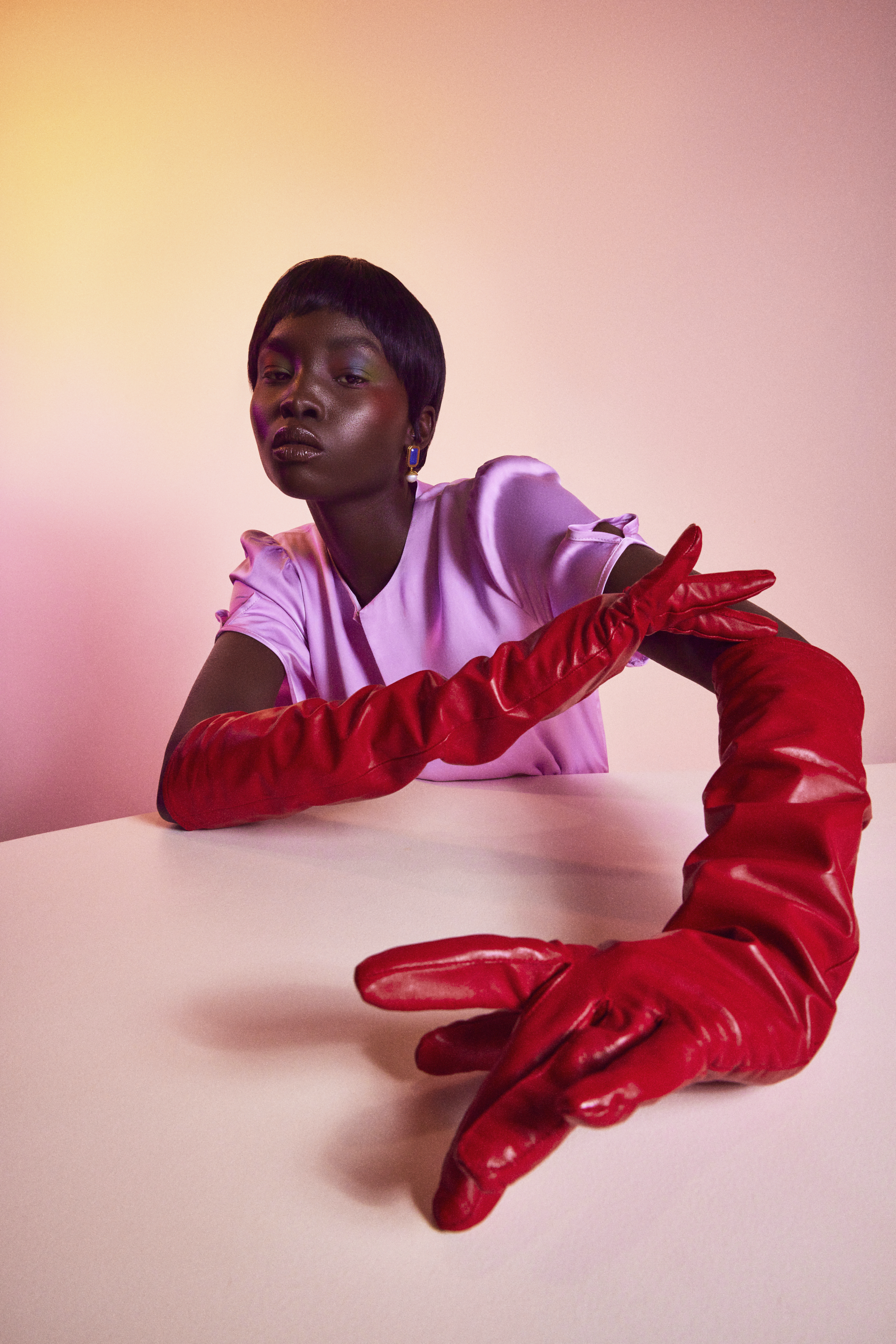 Black woman wearing long red leather gloves and a light purple top against a gradient pink backdrop by Lucy Alcorn
