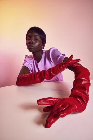 Black woman wearing long red leather gloves and a light purple top against a gradient pink backdrop by Lucy Alcorn