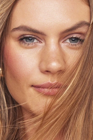 Close-up beauty portrait of a woman with glowing skin, pink lips, and golden blonde hair swept across her face.