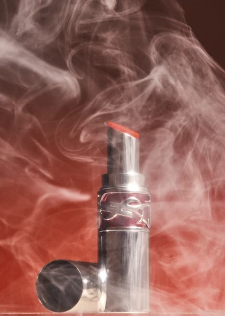 Silver lipstick tube with a vibrant red tip surrounded by swirling white smoke against a warm red background.