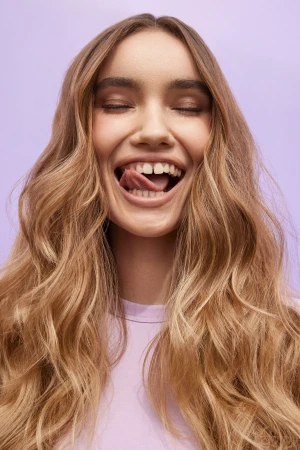 Close-up portrait of a woman with long wavy blonde hair sticking out her tongue against a soft purple background.