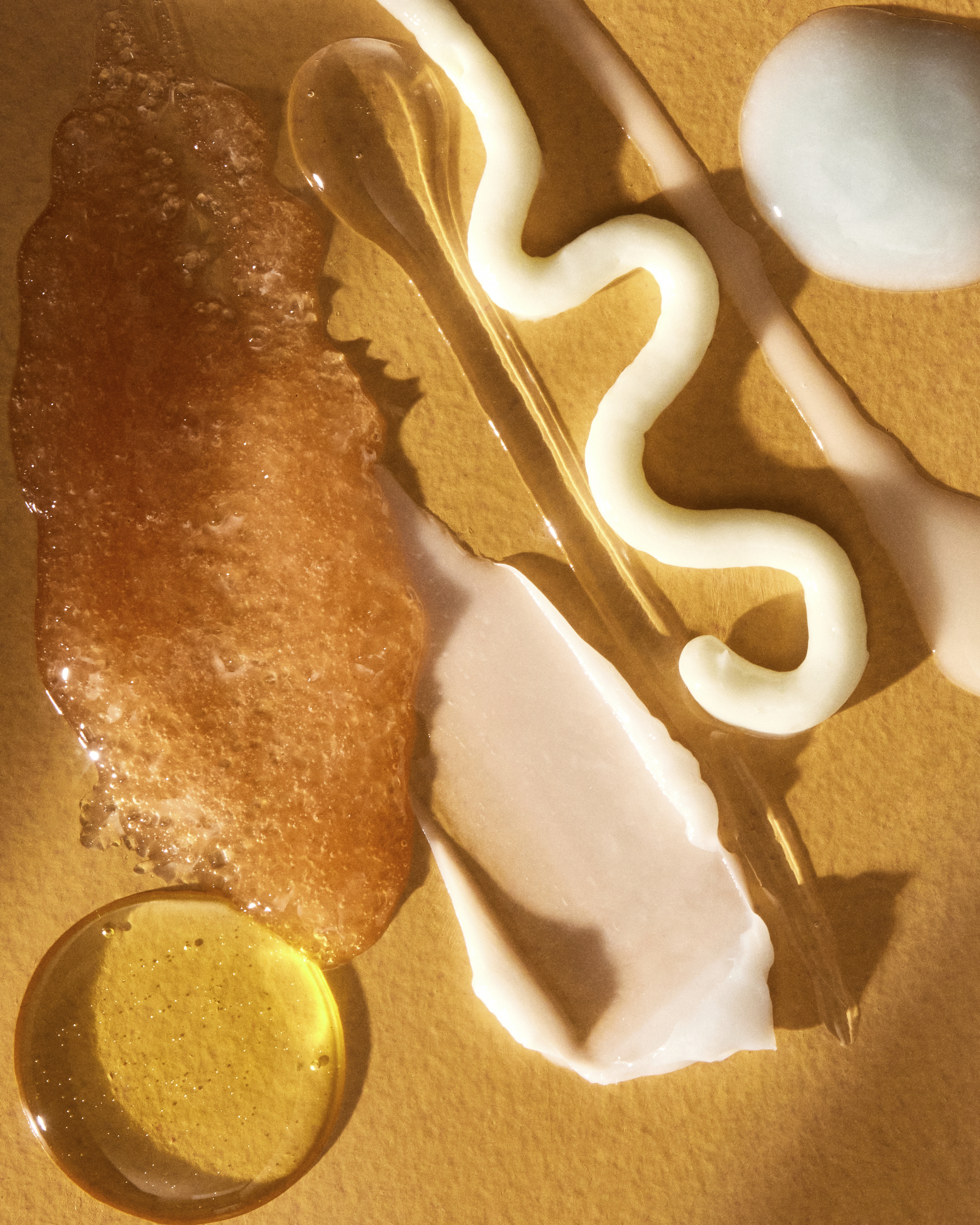 Close-up of skincare swatches and textures including creams, oil droplets, and scrub against a warm amber surface.